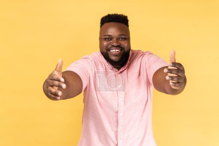 Photo for Come into my arms. Portrait of friendly man in pink shirt giving free hugs with outstretched hands, welcoming inviting to embrace, support and care. Indoor studio shot isolated on yellow background. - Royalty Free Image
