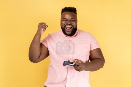 Photo for Portrait of extremely happy young adult man wearing pink shirt yelling happily, celebrating completing level in video games, holding joypad. Indoor studio shot isolated on yellow background. - Royalty Free Image