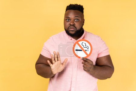 Photo for Portrait of young adult bearded man wearing pink shirt holding no smoking sign, showing stop gesture with palm, looking at camera. Indoor studio shot isolated on yellow background. - Royalty Free Image