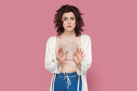 Photo for Portrait of scared shocked woman with curly hair wearing casual style outfit showing stop gesture, sees something frighten, looking at camera. Indoor studio shot isolated on pink background. - Royalty Free Image