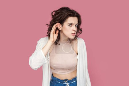 Photo for Portrait of serious curious woman with curly hair wearing casual style outfit keeps hand near ear, listening to something private, gossip. Indoor studio shot isolated on pink background. - Royalty Free Image