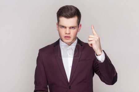 Photo for Young man being stern, raises fore finger up, frowns face, tries to attract attention to something very important, wearing violet suit and white shirt. Indoor studio shot isolated on grey background. - Royalty Free Image