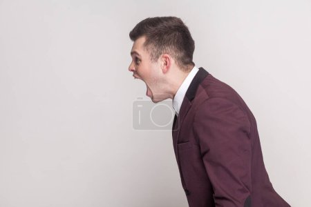 Photo for Side view portrait of aggressive annoyed handsome young man arguing with somebody, screaming with hate and anger, wearing violet suit and white shirt. Indoor studio shot isolated on grey background. - Royalty Free Image