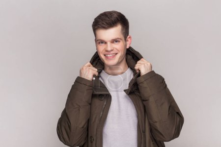 Photo for Portrait of optimistic young handsome man in light grey shirt and dark green parka, keeps hand on his collar, looking smiling at camera. Indoor studio shot isolated on gray background. - Royalty Free Image