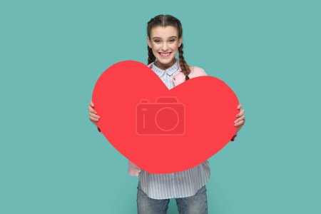 Photo for Portrait of happy joyful cheerful teenager girl with braids wearing pink jacket holding big red heart, looking at camera. Indoor studio shot isolated on green background. - Royalty Free Image