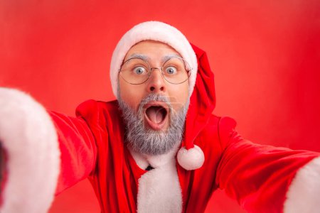 Téléchargez les photos : Portrait d'homme âgé avec barbe grise en costume de Père Noël faisant selfie POV ou diffusion en direct de la fête de Noël, expression excitée. Studio intérieur tourné isolé sur fond rouge. - en image libre de droit