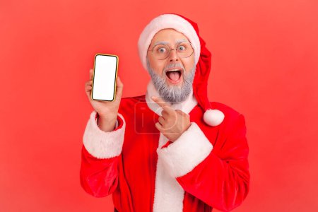 Photo for Portrait of amazed elderly man with gray beard wearing santa claus costume pointing at smart phone screen with copy space for ad, keeps mouth open. Indoor studio shot isolated on red background. - Royalty Free Image