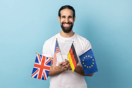 Téléchargez les photos : Portrait d'un homme extrêmement heureux avec une barbe portant un T-shirt blanc tenant dans les mains des drapeaux britanniques, allemands, européens et américains. Studio intérieur tourné isolé sur fond bleu. - en image libre de droit