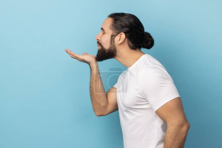 Photo for Side view portrait of romantic man with beard wearing white T-shirt blowing air kiss, folded palm towards mouth, sends mwah coquettish. Indoor studio shot isolated on blue background. - Royalty Free Image