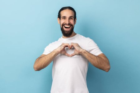 Photo for I love you sign. Be my valentine. Portrait of romantic man with beard wearing white T-shirt shapes heart confesses in love, looking at camera. Indoor studio shot isolated on blue background. - Royalty Free Image