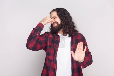 Photo for Ugh, what disgusting smell. Discontent bearded man with long curly hair in checkered red shirt covers nose, shows stop sign, notices rotten products. Indoor studio shot isolated on gray background. - Royalty Free Image