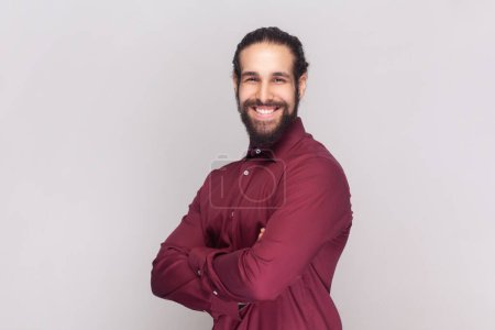 Photo for Portrait of smiling satisfied delighted man with dark hair and beard in red shirt standing with crossed arms, looking at camera with toothy smile. Indoor studio shot isolated on gray background. - Royalty Free Image