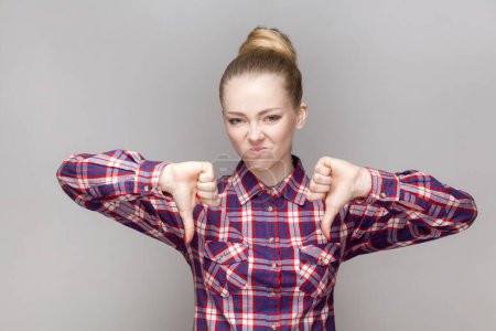 Photo for Portrait of disappointed sad woman with bun hairstyle standing with frowning face, showing dislike gesture, negative feedback, wearing checkered shirt. Indoor studio shot isolated on gray background. - Royalty Free Image