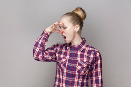 Photo for Portrait of disgusted adorable woman with bun hairstyle pinching her nose, turning away, feeling bad smell, wearing checkered shirt. Indoor studio shot isolated on gray background. - Royalty Free Image