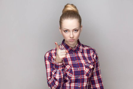 Photo for Portrait of strict bossy serious woman with bun hairstyle standing with raised finger, warning about dangerous situation, wearing checkered shirt. Indoor studio shot isolated on gray background. - Royalty Free Image
