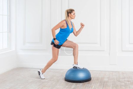 Photo for Side view of concentrated sporty beautiful young athletic woman wearing black shorts and blue top working in gym doing exercise in bosu balance trainer, lunges on fitness ball. Indoor studio shot. - Royalty Free Image