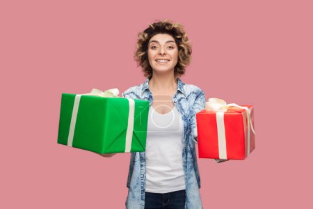 Photo for Smiling happy delighted woman with curly hairstyle wearing blue shirt standing giving two wrapped present box, congratulating her kids with holiday. Indoor studio shot isolated on pink background. - Royalty Free Image