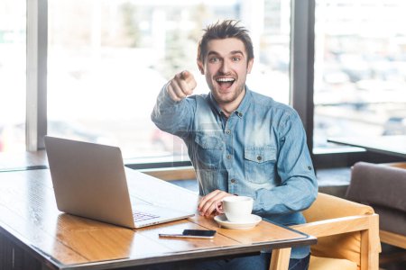 Téléchargez les photos : Portrait de jeune homme souriant et joyeux pigiste en jeans bleu chemise travaillant sur ordinateur portable, regardant pointant vers la caméra, vous choisissant. Plan intérieur près de la grande fenêtre, fond de café. - en image libre de droit