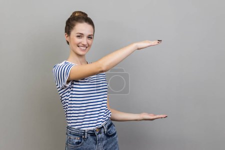 Photo for Portrait of happy smiling beautiful woman wearing striped T-shirt presenting area between hands for advertisement, showing huge size. Indoor studio shot isolated on gray background. - Royalty Free Image