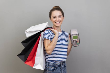 Photo for Portrait of satisfied woman wearing striped T-shirt holding payment terminal and paper shopping bags, easy express order and delivery. Indoor studio shot isolated on gray background. - Royalty Free Image