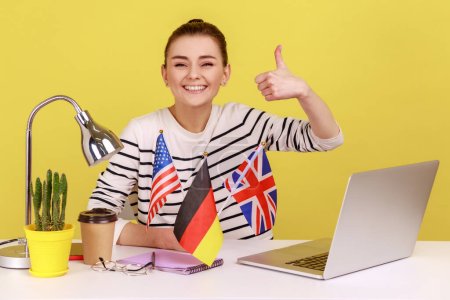 Téléchargez les photos : Satisfait femme souriante pointant vers les drapeaux américains, britanniques et allemands, se vantant de connaître de nombreuses langues, l'éducation à l'étranger. Studio intérieur tourné isolé sur fond jaune. - en image libre de droit