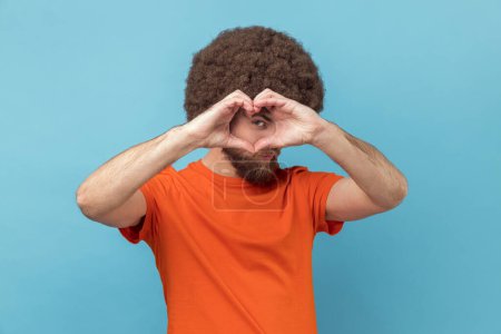 Téléchargez les photos : Portrait d'un bel homme romantique à la coiffure afro portant un T-shirt orange couvrant son visage d'un geste de cœur ou d'amour. Studio intérieur tourné isolé sur fond bleu. - en image libre de droit