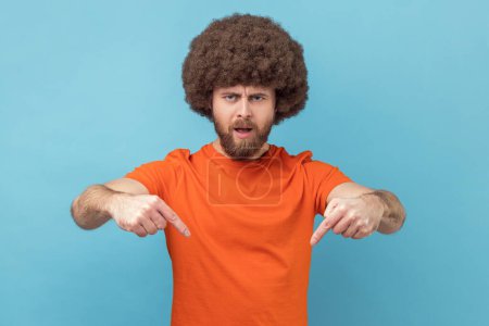 Photo for Portrait of man with Afro hairstyle wearing orange T-shirt pointing fingers down commanding to act here and now, demanding immediate result. Indoor studio shot isolated on blue background. - Royalty Free Image