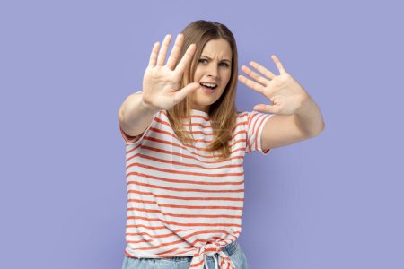 Photo for Portrait of afraid beautiful blond woman wearing striped T-shirt standing with raised arms, sees something awful, afraids, stop gesture. Indoor studio shot isolated on purple background. - Royalty Free Image