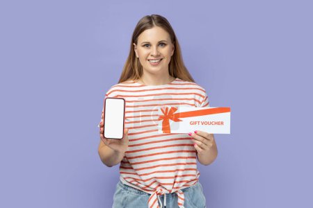 Photo for Portrait of attractive satisfied blond woman wearing striped T-shirt holding gift voucher and smart phone with empty display for promotion. Indoor studio shot isolated on purple background. - Royalty Free Image