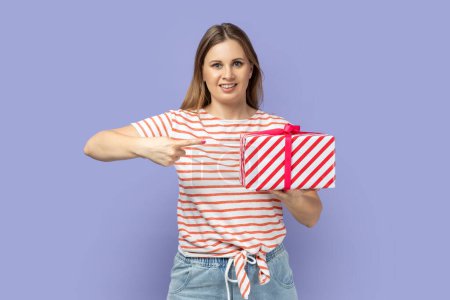 Photo for Portrait of pretty positive blond woman wearing T-shirt pointing to gift box with satisfied facial expression, looking at camera with toothy smile. Indoor studio shot isolated on purple background. - Royalty Free Image
