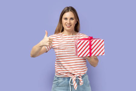 Téléchargez les photos : Portrait d'une femme heureuse et souriante portant un T-shirt rayé tenant un coffret cadeau et montrant le pouce levé, comme son cadeau, montrant un geste d'approbation. Studio intérieur tourné isolé sur fond violet. - en image libre de droit