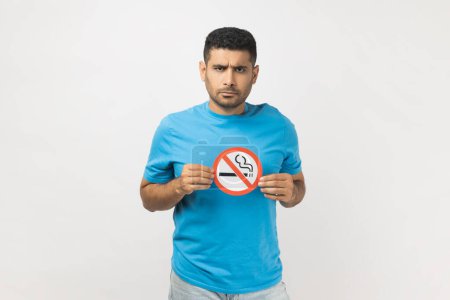 Photo for Portrait of strict serious handsome young adult unshaven man wearing blue T- shirt standing showing no smoking sign, looking at camera. Indoor studio shot isolated on gray background. - Royalty Free Image