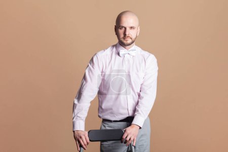 Photo for Portrait of representative handsome bearded man standing looking at camera with concentrated facial expression, wearing light pink shirt and bow tie. Indoor studio shot isolated on brown background. - Royalty Free Image