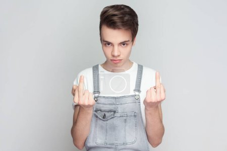 Photo for Portrait of rude impolite young brunette man standing shows middle finger bad sign, asking not to bother him, wearing denim overalls. Indoor studio shot isolated on gray background. - Royalty Free Image