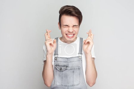 Photo for Portrait of happy young brunette man standing smiles broadly, keeps fingers crossed, hope for good luck during long trip, wearing denim overalls. Indoor studio shot isolated on gray background. - Royalty Free Image