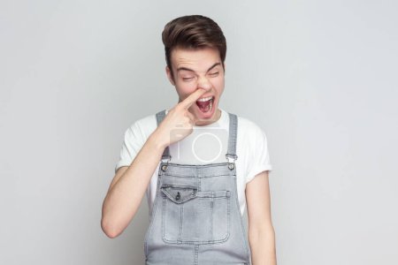 Photo for Portrait of impolite brunette man standing putting finger into his nose, fooling around, bad habits, disrespectful behavior, wearing denim overalls. Indoor studio shot isolated on gray background. - Royalty Free Image