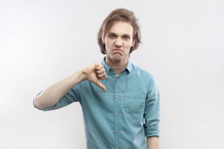 Photo for Portrait of young man criticizing bad quality with thumbs down displeased grimace, showing dislike gesture, expressing disapproval, wearing blue shirt. Indoor studio shot isolated on gray background. - Royalty Free Image
