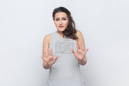 Photo for Oh no, stop it immediately. Portrait of brunette woman keeps palms forward, scared of something terrible, being afraid, wearing striped dress. Indoor studio shot isolated on gray background. - Royalty Free Image