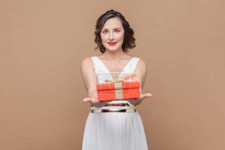 Photo for Portrait of optimistic middle aged woman with wavy hair holding giving red wrapped present box, congratulating with birthday, wearing white dress. Indoor studio shot isolated on light brown background - Royalty Free Image