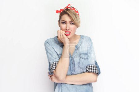 Photo for Portrait of sad nervous beautiful blonde woman wearing blue denim shirt and red headband standing biting her fingernails, having problems. Indoor studio shot isolated on gray background. - Royalty Free Image