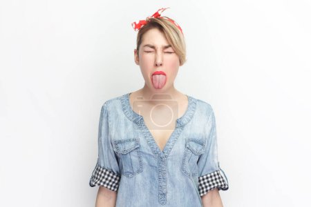 Photo for Portrait of funny foolish blonde woman wearing blue denim shirt and red headband standing with closed eyes, showing tongue out. Indoor studio shot isolated on gray background. - Royalty Free Image