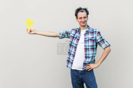 Photo for Smiling confident man holding little paper house in hand, real estate agency, accommodation, wearing blue checkered shirt and headband. Indoor studio shot isolated on gray background. - Royalty Free Image