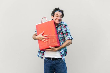 Photo for The best gift ever. Handsome man standing, hugging his lovely red shopping bag or gift and enjoying, wearing blue checkered shirt and headband. Indoor studio shot isolated on gray background. - Royalty Free Image