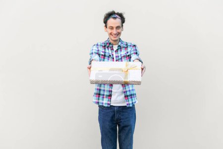 Photo for Satisfied man holding out gift box, congratulating friend with holiday, expressing positive emotions, wearing blue checkered shirt and headband. Indoor studio shot isolated on gray background. - Royalty Free Image