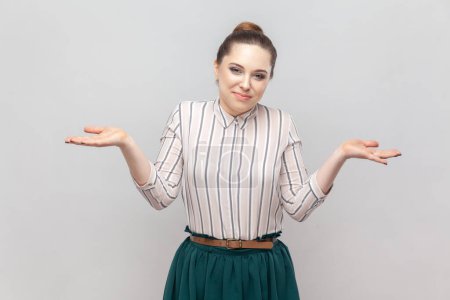 Photo for Portrait of attractive helpless young adult woman wearing striped shirt and green skirt shrugging her shoulders, doesn't know what to do. Indoor studio shot isolated on gray background. - Royalty Free Image