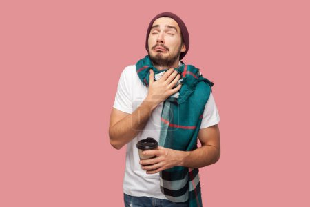 Photo for Portrait of sick handsome bearded man in white T-shirt, beany hat ans scarf standing with cup in hand, touching painful neck, suffering sore throat. Indoor studio shot isolated on pink background. - Royalty Free Image