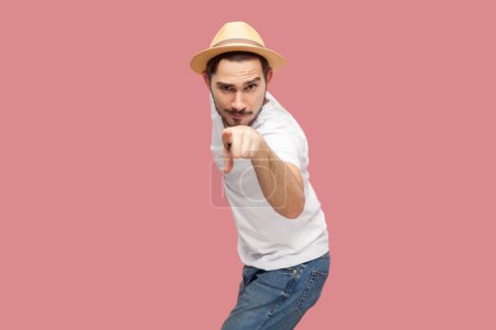 Téléchargez les photos : Portrait d'homme hipster barbu attrayant assertif sérieux et beau en T-shirt blanc et chapeau décontracté pointant vers la caméra, vous choisissant. Studio intérieur tourné isolé sur fond rose. - en image libre de droit