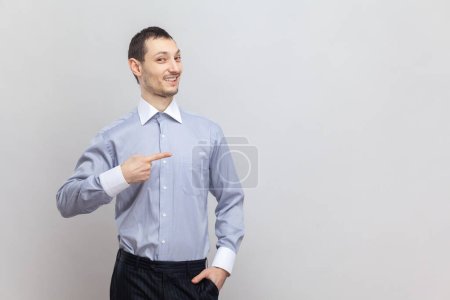 Téléchargez les photos : Portrait d'un homme souriant et optimiste, debout, pointant du doigt et faisant de la publicité, montrant l'espace de copie pour la promotion, portant une chemise bleu clair. Studio intérieur tourné isolé sur fond gris. - en image libre de droit
