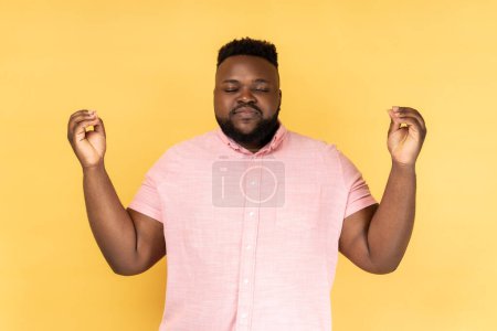Photo for Portrait of relaxed bearded man wearing pink shirt standing with raised arms and doing yoga meditating exercise, mudra gesture. Indoor studio shot isolated on yellow background. - Royalty Free Image