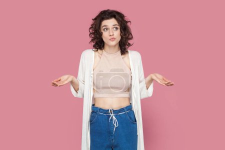 Photo for Portrait of pensive confused woman with curly hair wearing casual style outfit looking away with thoughtful expression, shrugging shoulders. Indoor studio shot isolated on pink background. - Royalty Free Image